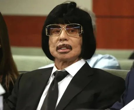 An Asian man with a black bowl-cut wig, mustache, and large tinted glasses wearing a black suit, white shirt, and black tie.