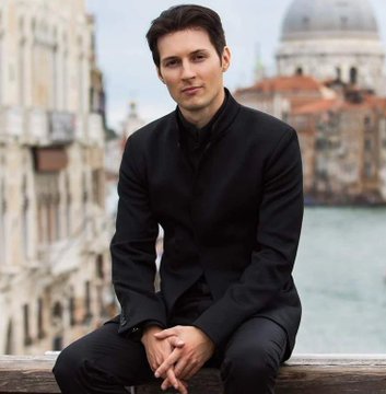 The image features a man sitting on a wooden bench or railing with his hands clasped together. He appears to be dressed in a black suit, which contrasts with the surrounding environment. The background includes buildings and water, suggesting that he might be in an urban setting near a body of water such as a river or canal. The post about crypto seems unrelated to the man's presence and attire, but it does capture attention due to its unusual title and content.