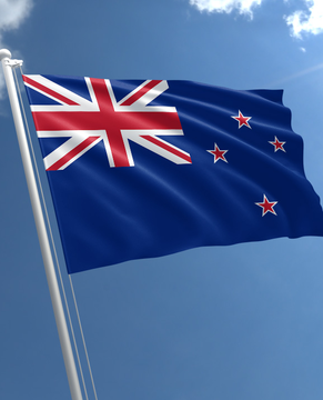 The image shows the flag of New Zealand, featuring the Union Jack in the upper left corner and four red stars with white borders on a blue background, waving against a clear blue sky. This flag visually represents New Zealand, the country whose 4th largest political party has pledged zero tax on Bitcoin and crypto assets.