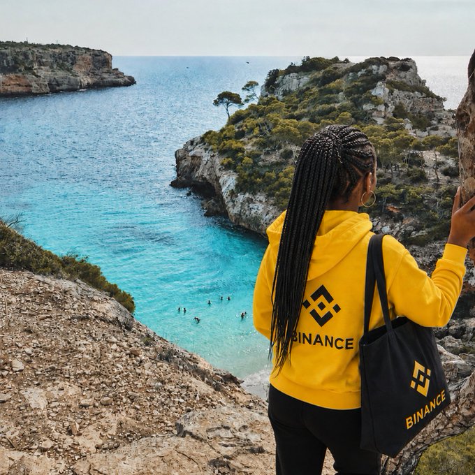 A woman in a yellow Binance hoodie and matching branded tote bag stands overlooking a stunning turquoise cove with swimmers below, embracing the peaceful coastal view. The serene seascape perfectly complements the message to slow down and savor life's simple moments.