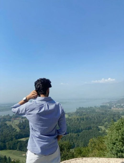 A man in a blue shirt standing with his back to the camera, looking out over a scenic lake and green landscape under a clear blue sky.