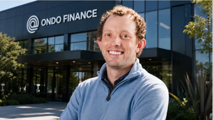 The image features a man standing outside of an office building, smiling for the camera. The man has short hair and is wearing a blue sweater. In front of him, there are two potted plants that add some greenery to the scene. The background includes a large sign with the words "ONDO Finance" written on it in white letters.