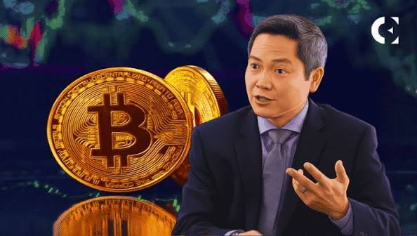 A man in a dark suit and purple tie speaks in an interview setting, gesturing with his hand, with large golden Bitcoin coins displayed behind him on a screen.