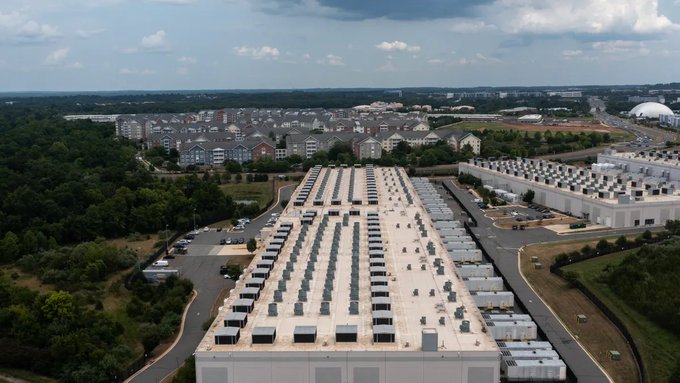 An aerial view of Amazon's expansive data center complex with rows of cooling units on rooftops, set against a backdrop of residential buildings and a cloudy sky, illustrating the vulnerability of centralized cloud infrastructure to physical attacks—as highlighted by the post about Iranian strikes on data hubs and the need for decentralized alternatives like Dfinity's ICP cloud engines.