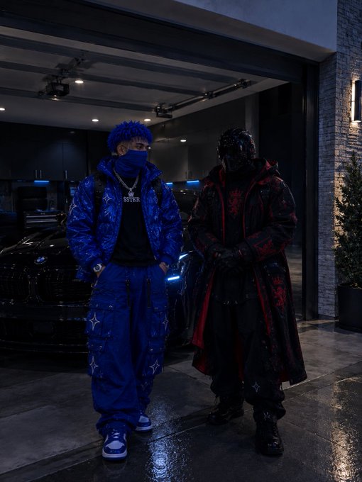 Two figures in avant-garde streetwear stand in front of a black luxury car in a garage setting; one wears an all-blue monogrammed outfit with a matching face covering, and the other wears a black and red monogrammed outfit with a dark mask.
