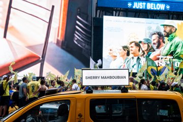 A yellow taxi with a "SHERRY MAHBOUBIAN" nameplate displays the artist's work "To Belong" on a digital rooftop screen in a busy urban setting, with a "JUST BELOW" advertisement visible on a building behind it, bringing her featured exhibition to the streets of New York.
