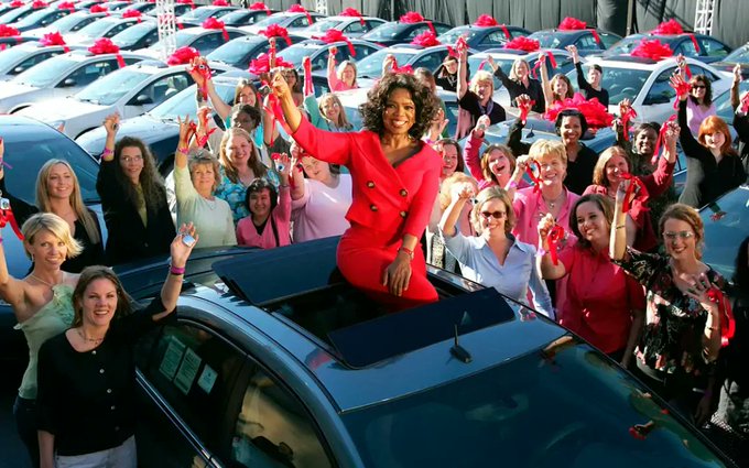 The image shows a large group of people standing around cars, with some women wearing red dresses holding up their arms high above their heads. The scene appears to be at a car dealership or an event related to automobiles. The caption suggests that there is a competition for who can fill out the most forms correctly in the image.