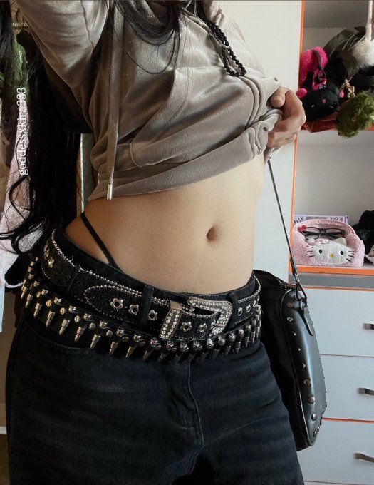 A close-up photo of a person's torso wearing a cropped gray hoodie, black jeans, and a black studded belt, with what appears to be a thigh holster strapped to their leg.