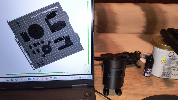 A workspace setup showing a laptop screen displaying 3D models of black plastic parts on a grid-patterned print bed (appearing to be Bambu Lab 3D printer components), alongside a small black cylindrical device, a "SAC" brand bottle, and a "Lovia" nutritional supplement container on the desk.