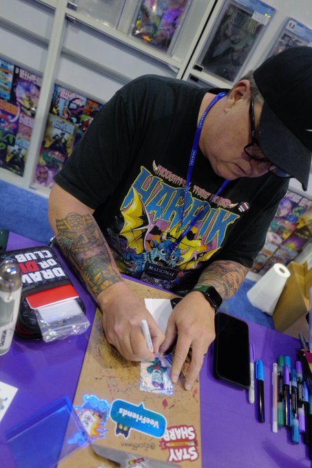 A tattooed man in a black cap and 'SHARPIE STARBOX' t-shirt sketches on a card at a comic convention table.