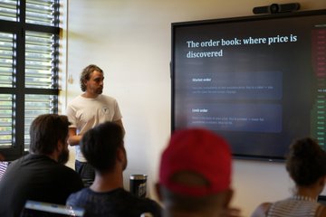 A presenter in a white shirt stands before a screen displaying "The order book: where price is discovered," while an audience watches attentively, capturing the educational and collaborative atmosphere of the month-end recap within the RGB ecosystem.