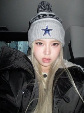 A young woman with long blonde hair wears a gray star-patterned beanie with a pom and a dark winter coat.