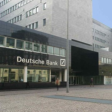 The image shows the exterior of a Deutsche Bank building, identifiable by the company's name and logo displayed prominently above the entrance, along with a few people walking in the plaza area outside. This connects to the news about Deutsche Bank's plans to offer regulated Bitcoin and Ether custody services for institutional clients in Europe by end-2026, highlighting the bank's expansion into cryptocurrency services.