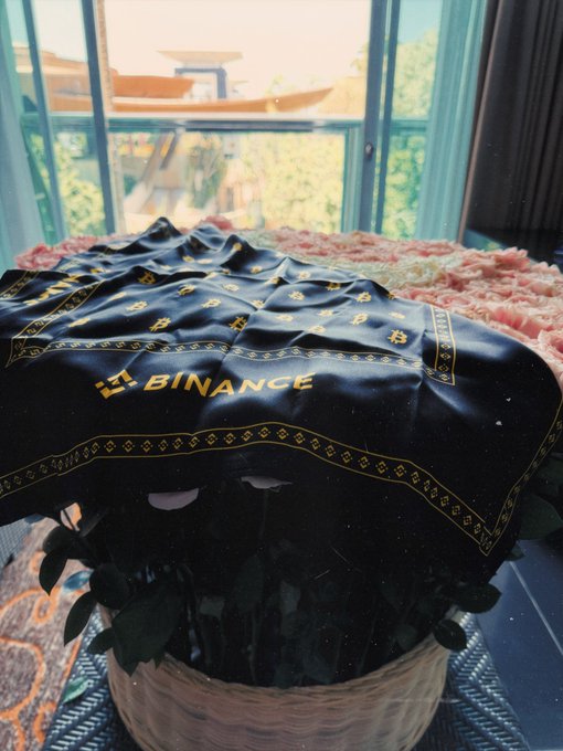 A bouquet of pink roses is wrapped in black paper prominently featuring the yellow "BINANCE" logo and crypto-style patterns.
