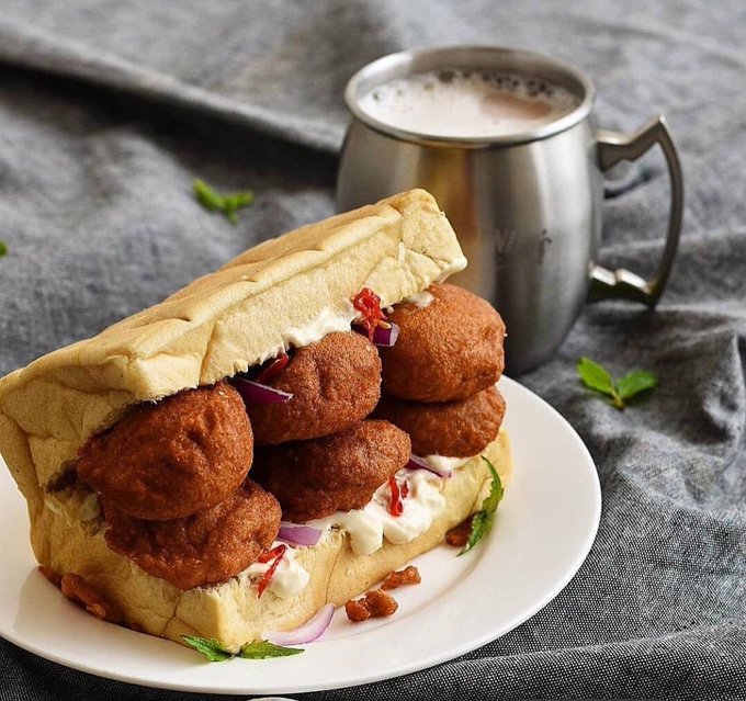 A close-up photo of a sub sandwich filled with fried meatballs, red onions, and white sauce, served on a white plate alongside a metal mug of coffee.