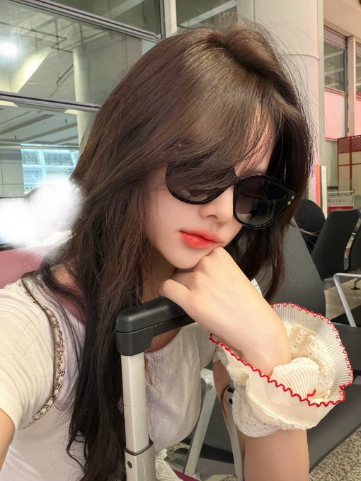 Young woman with long brown hair wearing dark sunglasses and a white ruffled top, posing with a suitcase handle in what appears to be an airport terminal.