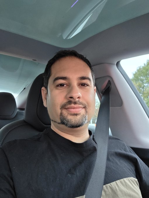 A man takes a selfie while sitting in the driver's seat of a car, wearing a black shirt and a seatbelt.