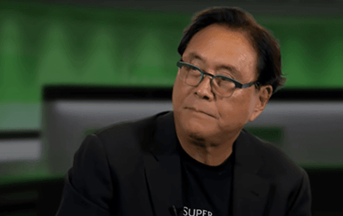 Close-up shot of an older Asian man wearing round glasses and a black blazer over a t-shirt, looking to the side with a slight smirk against a green background.