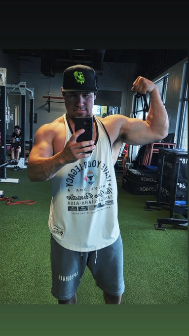A muscular man in a black cap and white tank top takes a mirror selfie while flexing his bicep in a gym.