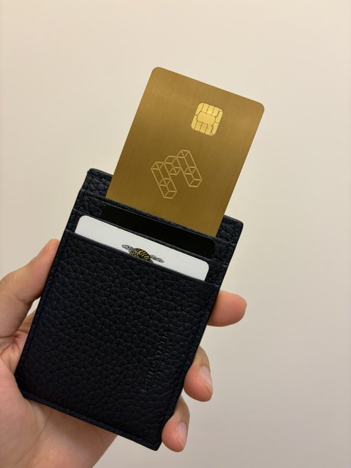 A hand holds a black leather wallet containing a gold card featuring an EMV chip and a geometric logo, alongside a white card partially visible with a bee emblem. The luxurious gold card aligns with the post's flex of an EtherFi crypto-related card, showcasing a premium financial product.