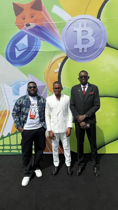 Three men pose in front of a colorful banner featuring a Shiba Inu dog, a Bitcoin (₿) coin, and other crypto-related graphics, with one man wearing a "BNB" branded t-shirt.