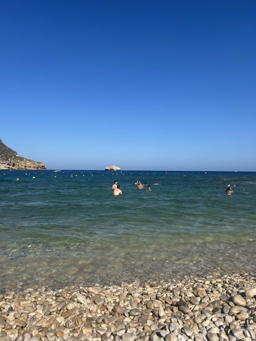 A group of people enjoy a final swim in the crystal-clear turquoise waters of a rocky beach, with a small island visible in the distance under a bright blue sky. The serene seaside scene captures a peaceful last day of vacation before returning to trading and content creation.