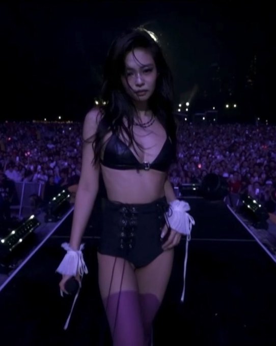 A female performer in a black leather crop top, lace-up skirt, and purple thigh-high stockings walks on a stage during a concert, with a large crowd visible in the background.