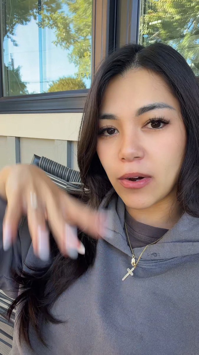 A selfie of a young woman wearing a gray hoodie and a cross necklace, outdoors with her hand reaching toward the camera.