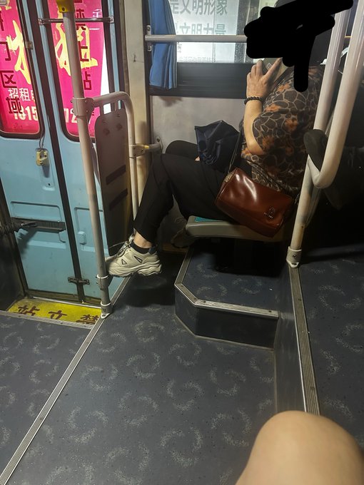 The image shows a woman sitting down, possibly on a bus or subway car, with her legs crossed. She has a purse next to her and appears to be looking at something inside the bag. The photo also includes Chinese text that reads "这就是福建！！ 刚上公交就听见阿姨讲 被套 割掉 今天行情不好玩". This text seems to be a reference or description of the woman's situation, possibly related to her travel experience.

The image also features a bus stop sign in the background and a person holding a cell phone. The presence of these elements suggests that this scene takes place at a transit hub where people are waiting for their transportation or just disembarking from one.