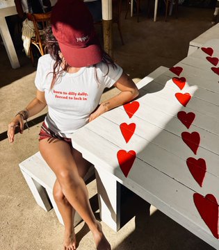 A woman wearing a red cap and white t-shirt reading 'born to defy rules, forced to back it' sitting on a white wooden bench decorated with red heart shapes.