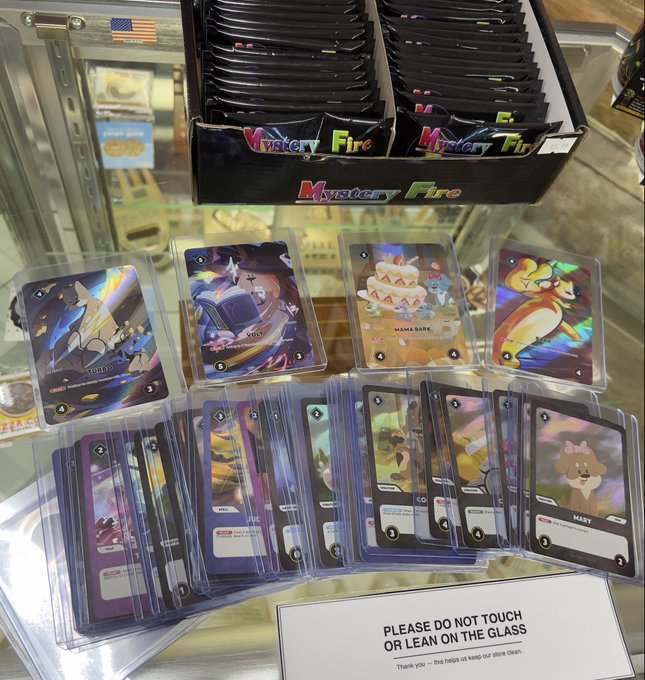 Holographic trading cards featuring characters like Garfield, Mama Bear, and others are displayed on glass alongside "Mystery Fire" packs, with a sign warning "PLEASE DO NOT TOUCH OR LEAN ON THE GLASS."