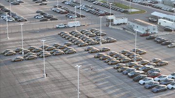 An aerial view shows a large parking lot filled with hundreds of what appear to be Cybercab robotaxis arranged in neat rows, supporting the post's claim of ramping production toward 10,000+ Cybercabs by December. The orderly fleet suggests significant manufacturing output, though no specific text or numbers are visible in the image itself.