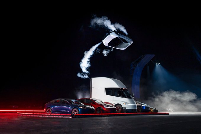 A Tesla Semi truck launches another vehicle into the air on a dramatic stage, illustrating the post's point about how cars and their stunts are increasingly priced in Bitcoin—featuring several Tesla models displayed on a smoky platform.