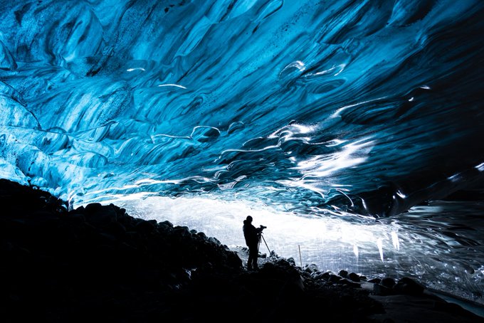A silhouette of a photographer standing inside a striking blue ice cave, aiming his camera at the illuminated, textured glacial wall.