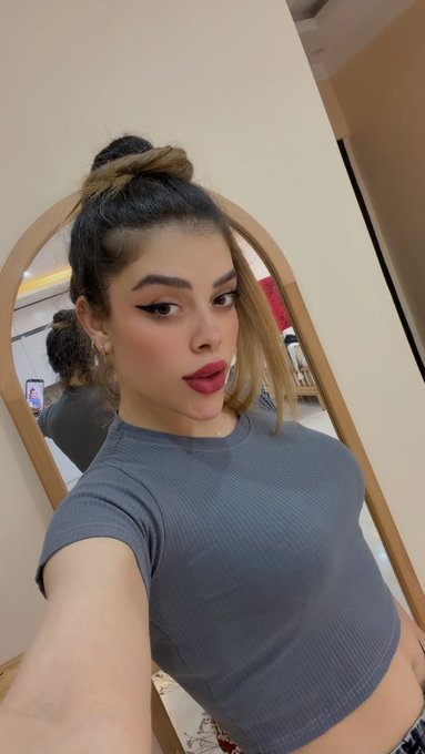A selfie of a young woman with her hair in a top knot and highlights, wearing a gray ribbed short-sleeve shirt, making a pouting face in front of a mirror.