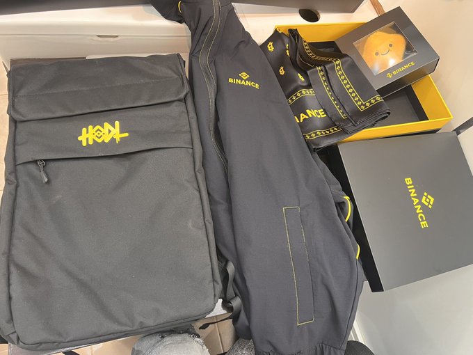 A collection of Binance-branded merchandise is displayed, featuring a backpack with "HODL" printed on it, a jacket, and a box containing a plush toy. The gifts, prominently displaying the "BINANCE" logo, appear to be appreciation swag from the cryptocurrency exchange.