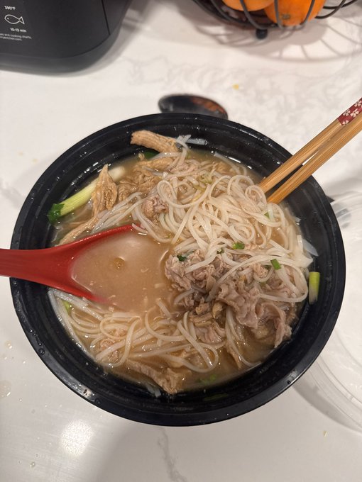 A black bowl filled with noodle soup containing meat and green vegetables is shown with a red spoon and chopsticks resting inside.