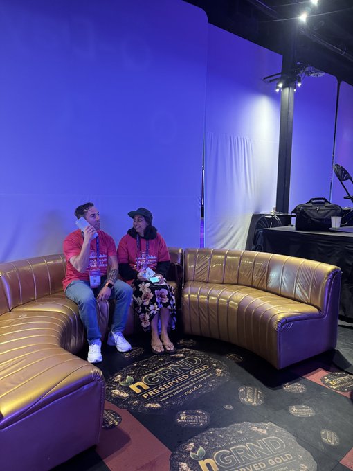 Two people sitting on a curved gold leather sofa in a purple-lit lounge area with 'nGRND Preserved Gold' branded rugs on the floor.