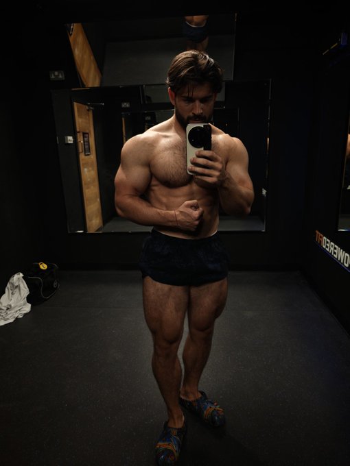 A shirtless, muscular man takes a mirror selfie in a gym locker room while flexing his arm, wearing only dark shorts and patterned sneakers. The text "POWERED" is visible on the wall, and the image humorously pairs with the post about a "pump" by showcasing his visibly pumped physique alongside a tongue-in-cheek reference to Bitcoin's price target.