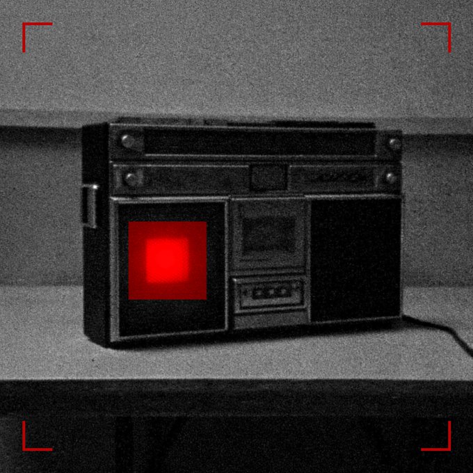 A black and white image of an old radio or cassette player with a glowing red light, framed by red corner brackets as if viewed through a viewfinder.
