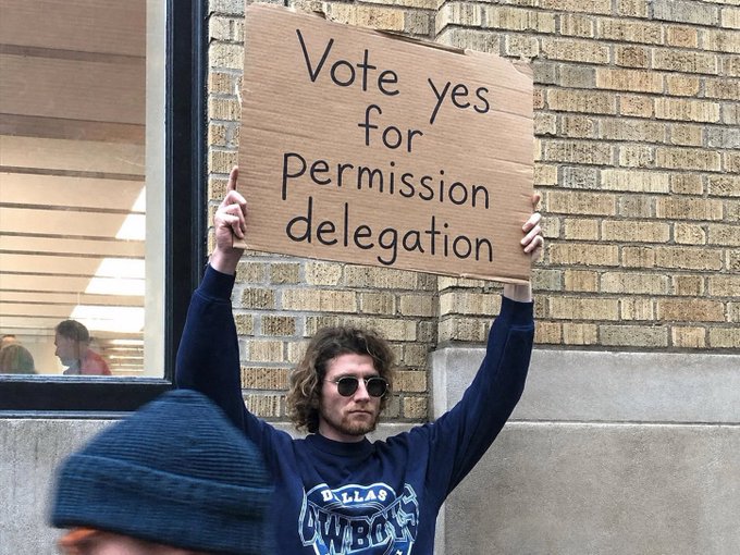 A young man with curly hair and sunglasses holds up a cardboard sign reading "Vote yes for Permission delegation" outside a brick building, supporting an XRPL amendment aimed at enabling a large asset manager to use the ledger. The sign's message directly aligns with the post's call for UNL validators to review and approve the amendment.