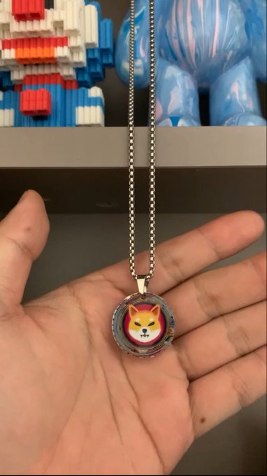 The image features a hand holding up a necklace with a small cartoon cat face on it, which has been designed by Shibin Chen. The cat's face is yellow with red eyes and a pink nose, giving it a unique and playful appearance. The necklace is made of metal and is displayed against the backdrop of a gray wall. The post about the cat necklace includes information such as its creator, Shibin Chen, and that it was created in 2008.