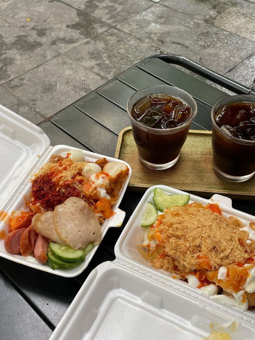 Two styrofoam takeout containers of food with rice, cucumber slices, sausage, fried toppings, and sauce, accompanied by two glasses of iced dark beverage on a wooden tray at an outdoor table.