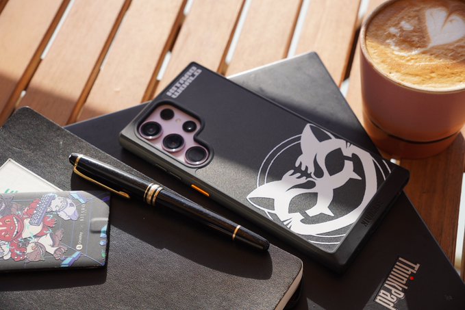 A close-up, sunlit photo of a black smartphone case with a silver wolf emblem, a black pen, and a notebook on a wooden table next to a cup of latte; the case appears to be a ThinkPad-branded collaboration.