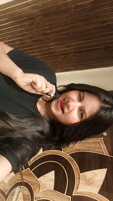 A woman taking a selfie with the image oriented sideways, smiling slightly with red lipstick and long dark hair, holding what appears to be a makeup product near her face. She seems to be saying "Gm" (good morning) back to the community with a casual, friendly selfie vibe.
