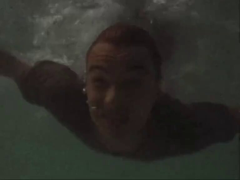 The image shows a person swimming underwater with their head above water, wearing a brown shirt. The surrounding water appears greenish-blue, suggesting that they are in an indoor pool or a body of water with a similar coloration. The photo is taken from below and slightly to the left, giving us a view of the person's face as well as their upper body. The image also contains text that reads "Onto a New Chapter, Arx Mode Active Praise the Sacred Parasol Mark it 6.22 Purplethon". This could be an advertisement or promotional material for a product related to swimming or underwater activities.