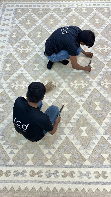 Two people in black t-shirts kneel on a patterned tile floor, using small tools to clean or work on the grout between the tiles.