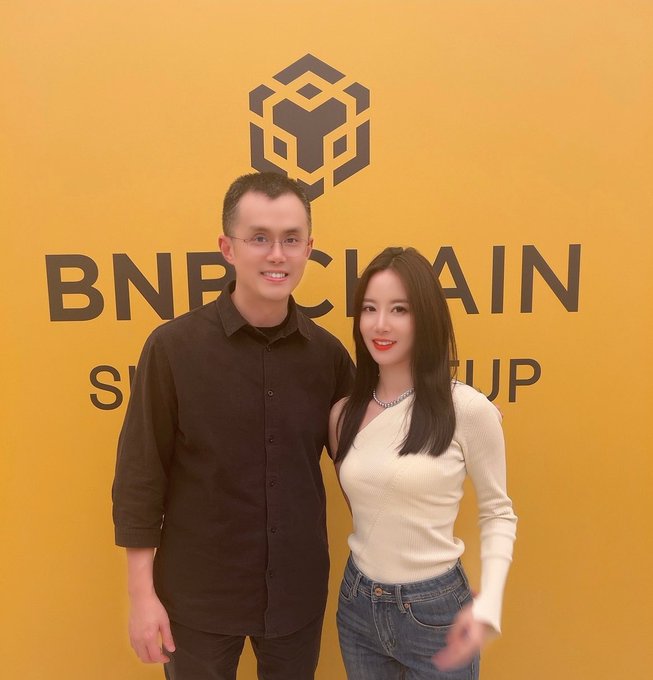Two people stand in front of a yellow backdrop featuring the BNB Chain logo and text reading 'BNB CHAIN SUMMIT'.