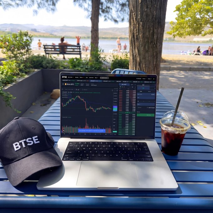 A trader wearing a BTSE cap works remotely by a scenic lake, analyzing Bitcoin price charts on their laptop alongside an iced coffee, capturing the "fully locked in" focus and fresh optimism of a new trading week.