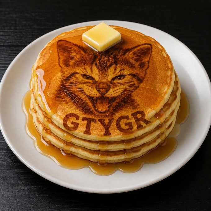 A stack of pancakes on a white plate with a pat of butter on top, featuring a pancake imprinted with a snarling cat face and the text 'GTYGR' below it, drizzled with syrup.